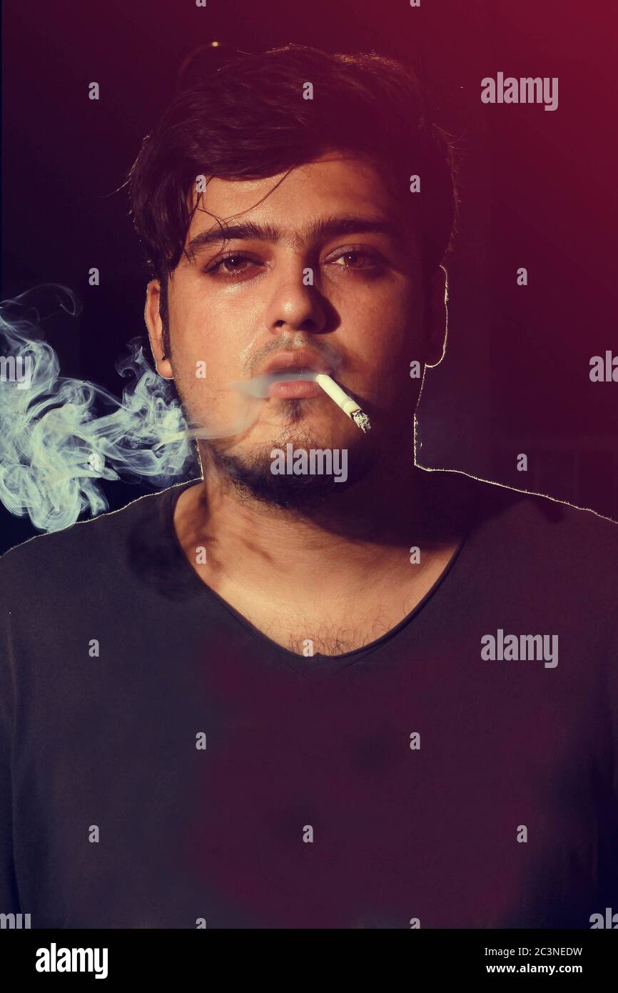 Young man smoking cigarette on dark background looking into camera wearing t shirt Stock Photo
