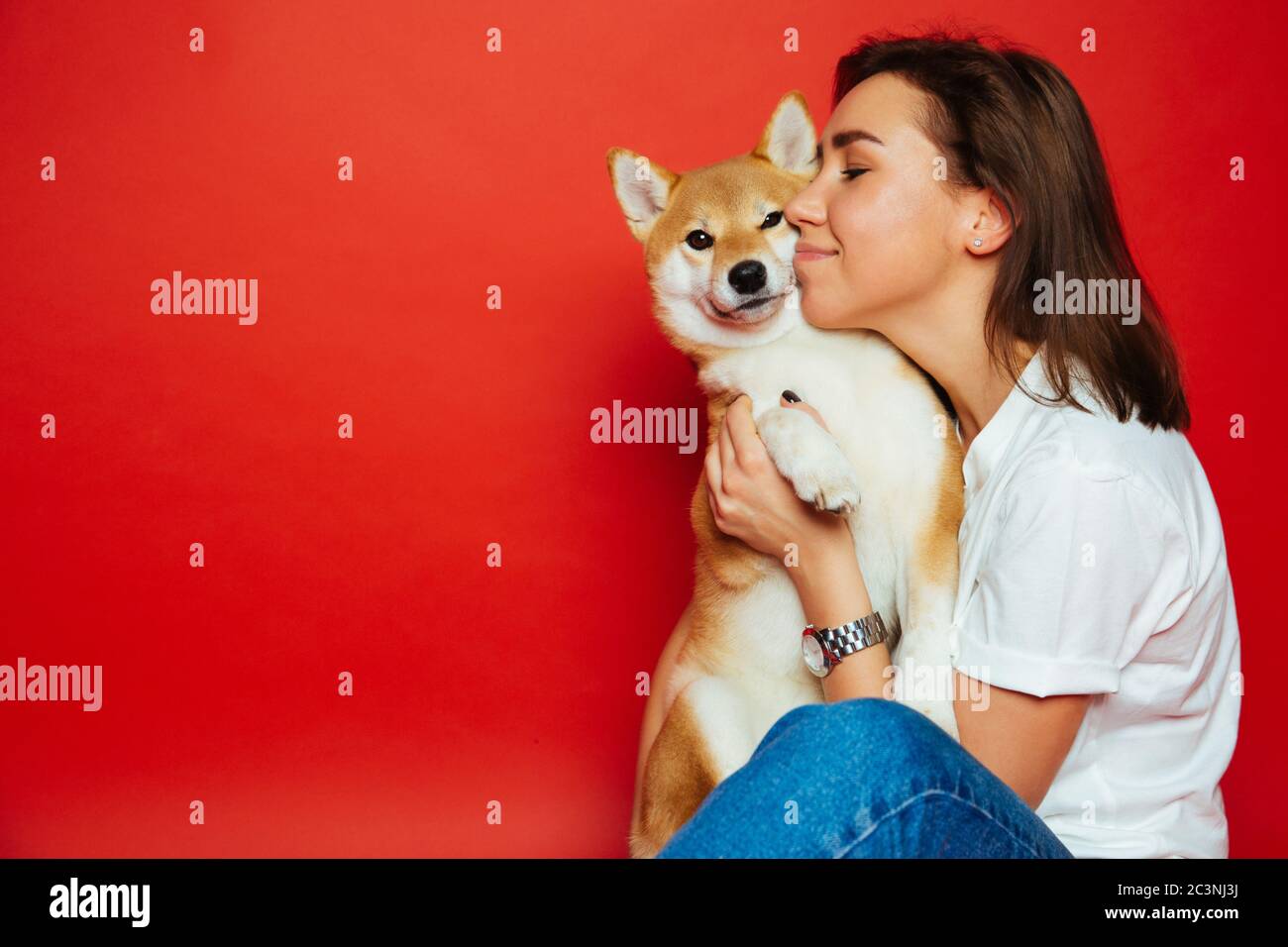 Cute brunette woman in white t shirt and jeans holding and embracing Shiba Inu dog on plane red background. Love to the animals, pets concept Stock Photo