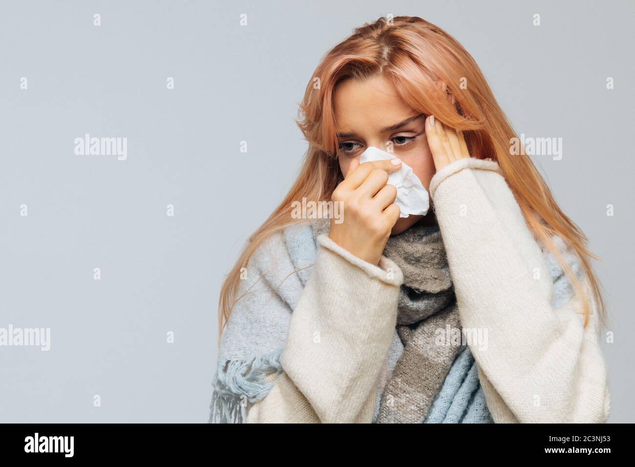 Attractive sick young woman with strawberry blonde hair wrapped in warm scarf with napkin blowing nose, feels bad, having headache, touching her foreh Stock Photo