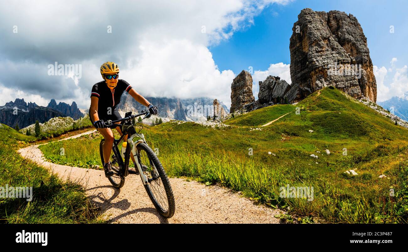 Tourist cycling in Cortina d'Ampezzo, stunning Cinque Torri and Tofana in background. Woman and man riding MTB trail. South Tyrol province of Italy, D Stock Photo