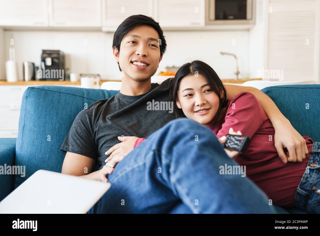 Photo of beautiful joyful asian couple watching tv and using remote control while sitting on sofa at home Stock Photo