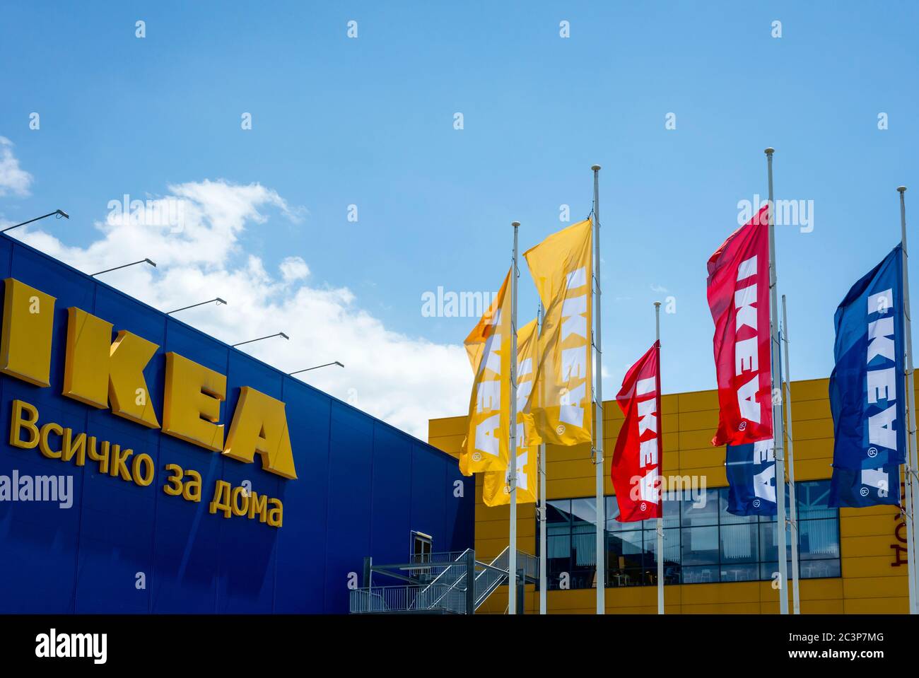 IKEA sign and brand logo in Cyrillic writing at the Sofia Bulgaria store branch with multicolored or multicoloured waving flags against blue sky Stock Photo