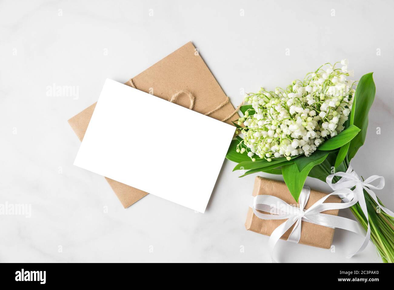 blank greeting card with lily of the valley flowers bouquet and gift box on white background. mock up. flat lay. top view. wedding invitation Stock Photo