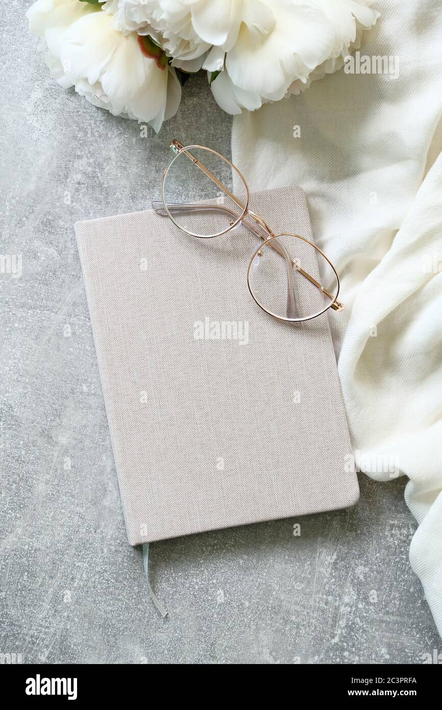 Flat lay composition with beige paper notebook, hipster glasses, textile cloth and peonies on concrete stone background. Elegant feminine workspace to Stock Photo