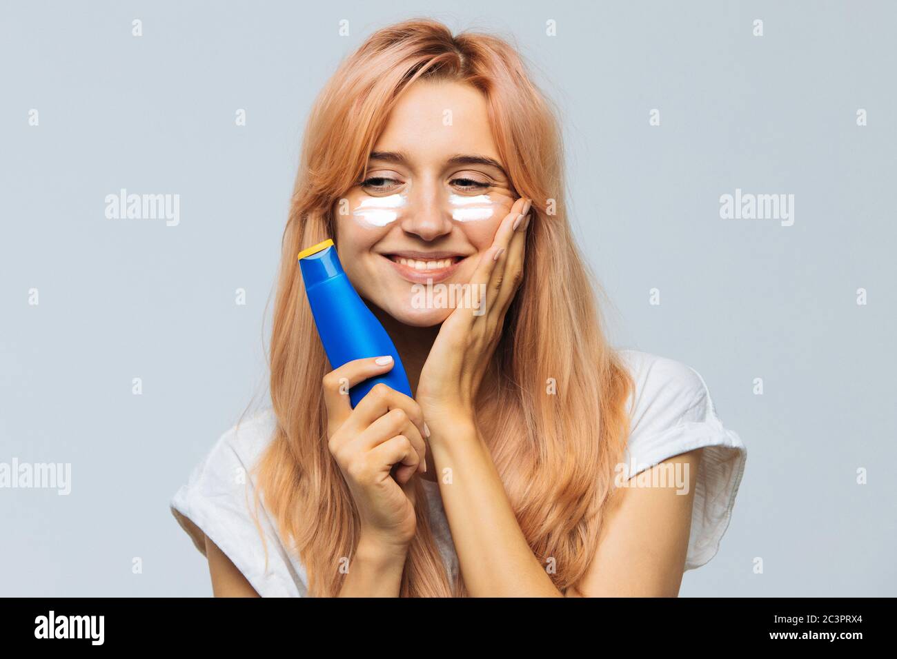 Portrait of young joyful cute woman smiling and applying suntan cream (sunscreen lotion) on face. Pretty female smeared face, cheeks with sun protecti Stock Photo