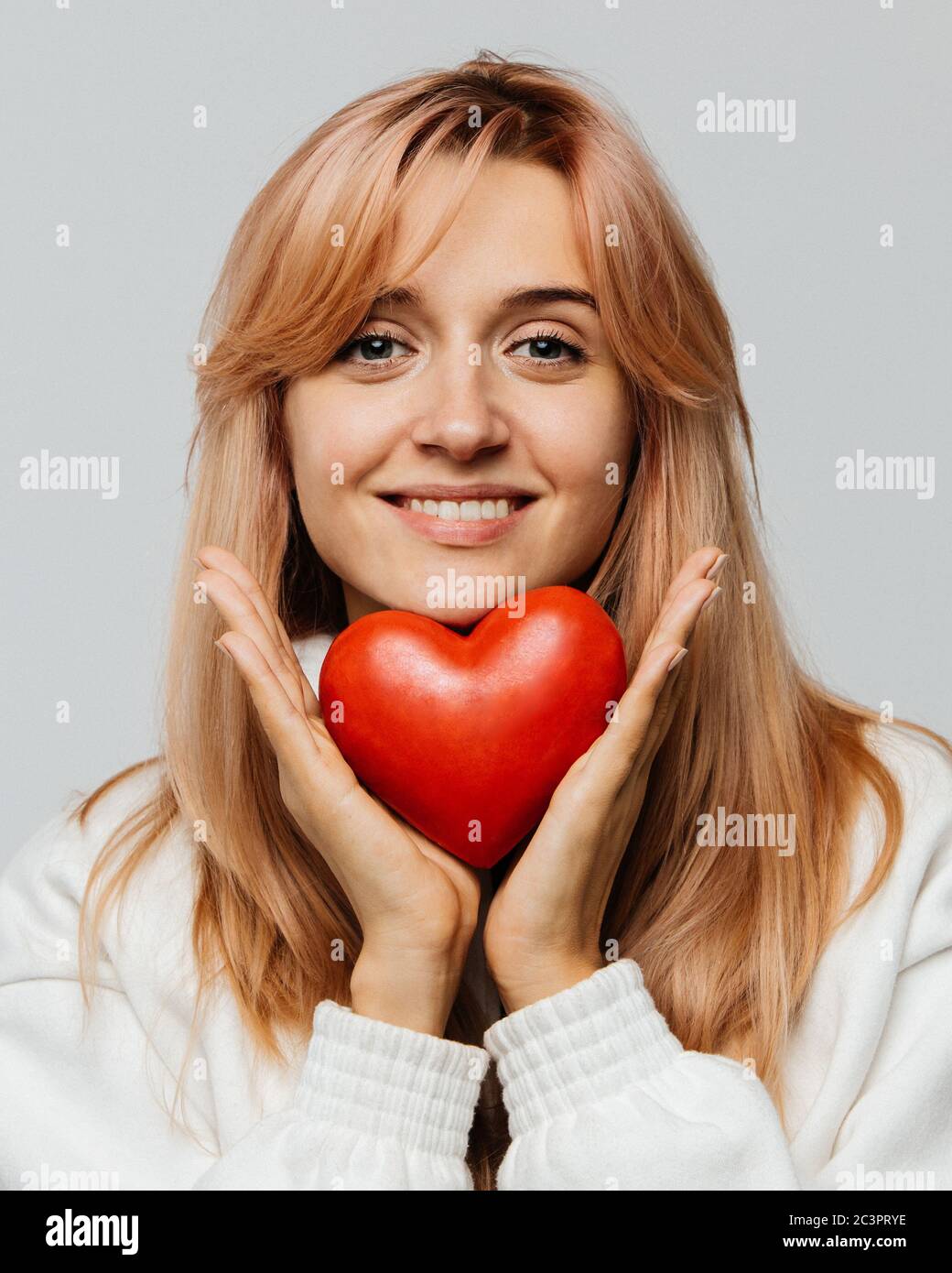 Portrait of joyful attractive woman with strawberry blonde hair hold red heart (Valentine day symbol),looking at camera, closeup, isolated on light ba Stock Photo