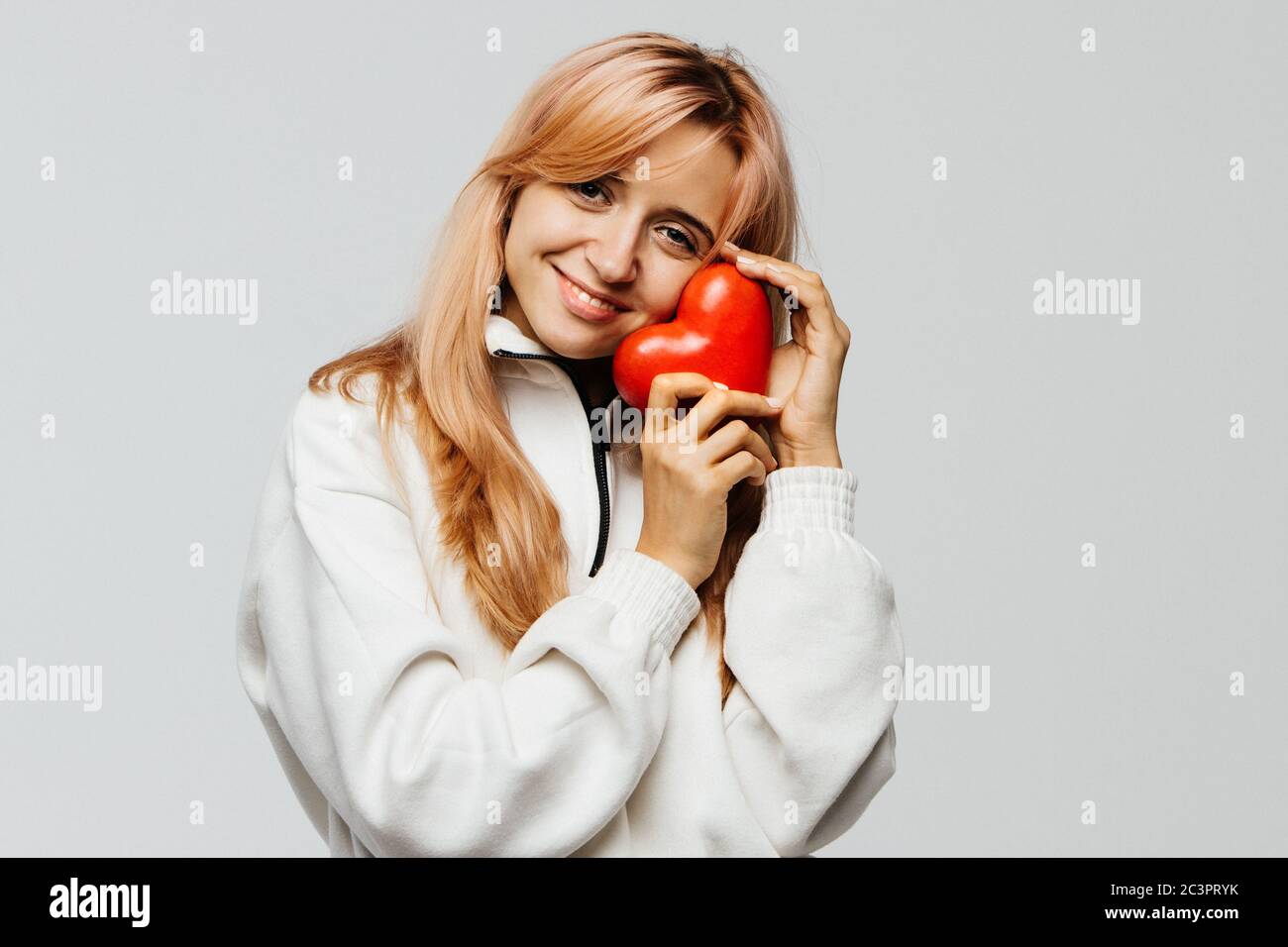 Portrait of joyful attractive woman with strawberry blonde hair hold red heart (Valentine day symbol),looking at camera, closeup, isolated on light ba Stock Photo