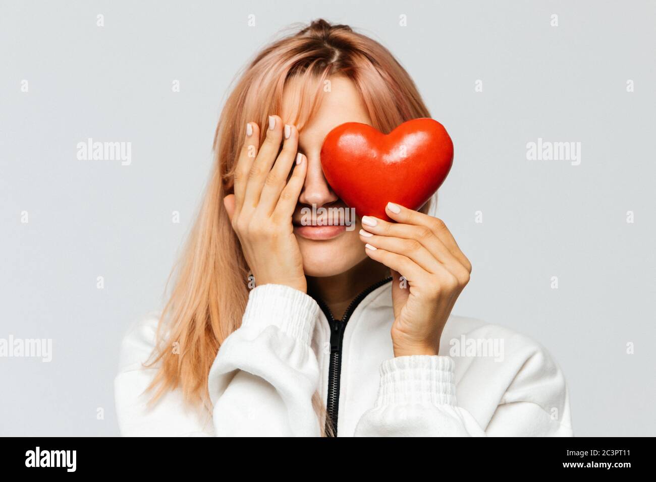 Happy cute young woman with strawberry blonde hair hold red heart (Valentine day symbol),covering her right eye with her palm, closeup, isolated on li Stock Photo