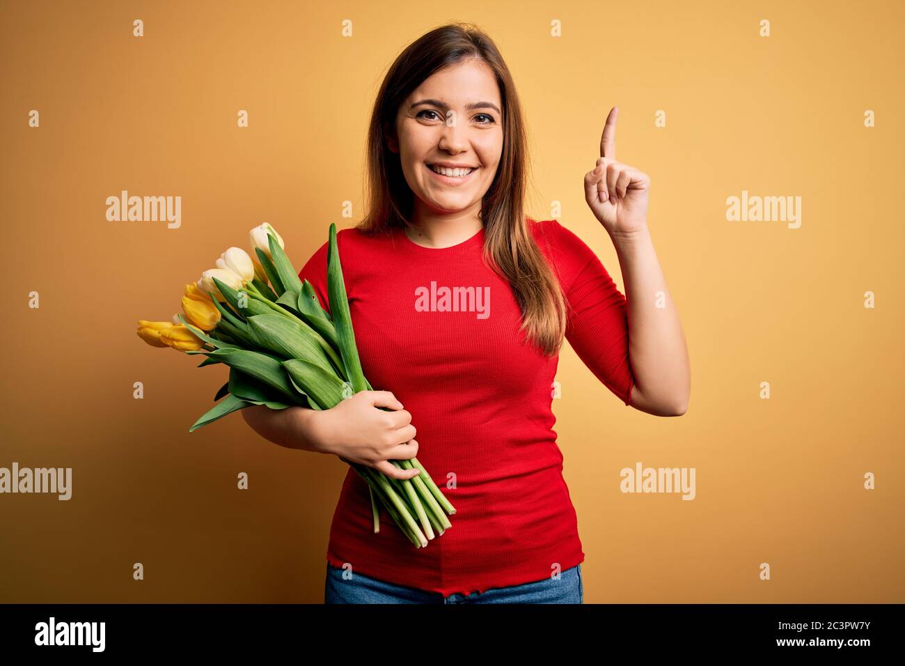 Young blonde woman holding romantic bouquet of tulips flowers over yellow background smiling amazed and surprised and pointing up with fingers and rai Stock Photo