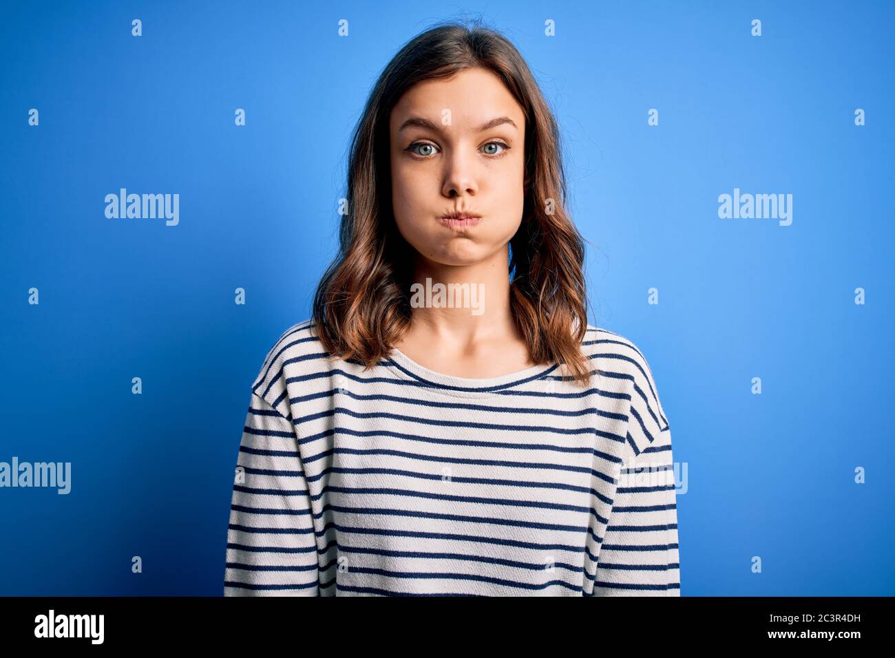 Young beautiful blonde girl wearing casual sweater standing over blue isolated background puffing cheeks with funny face. Mouth inflated with air, cra Stock Photo