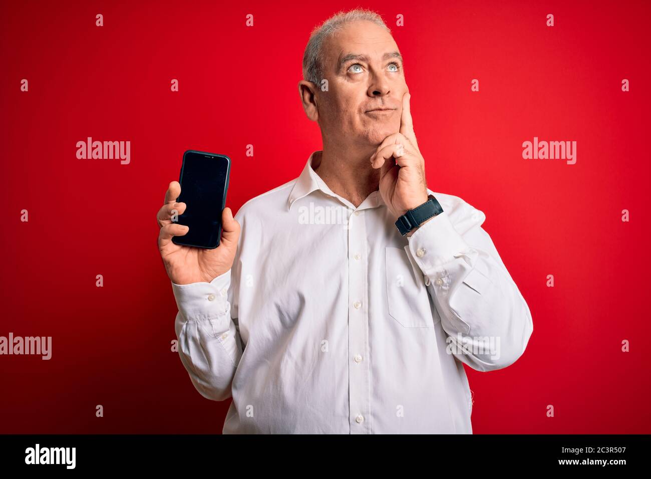 Middle age hoary man holding smartphone showing screen over isolated red background serious face thinking about question, very confused idea Stock Photo