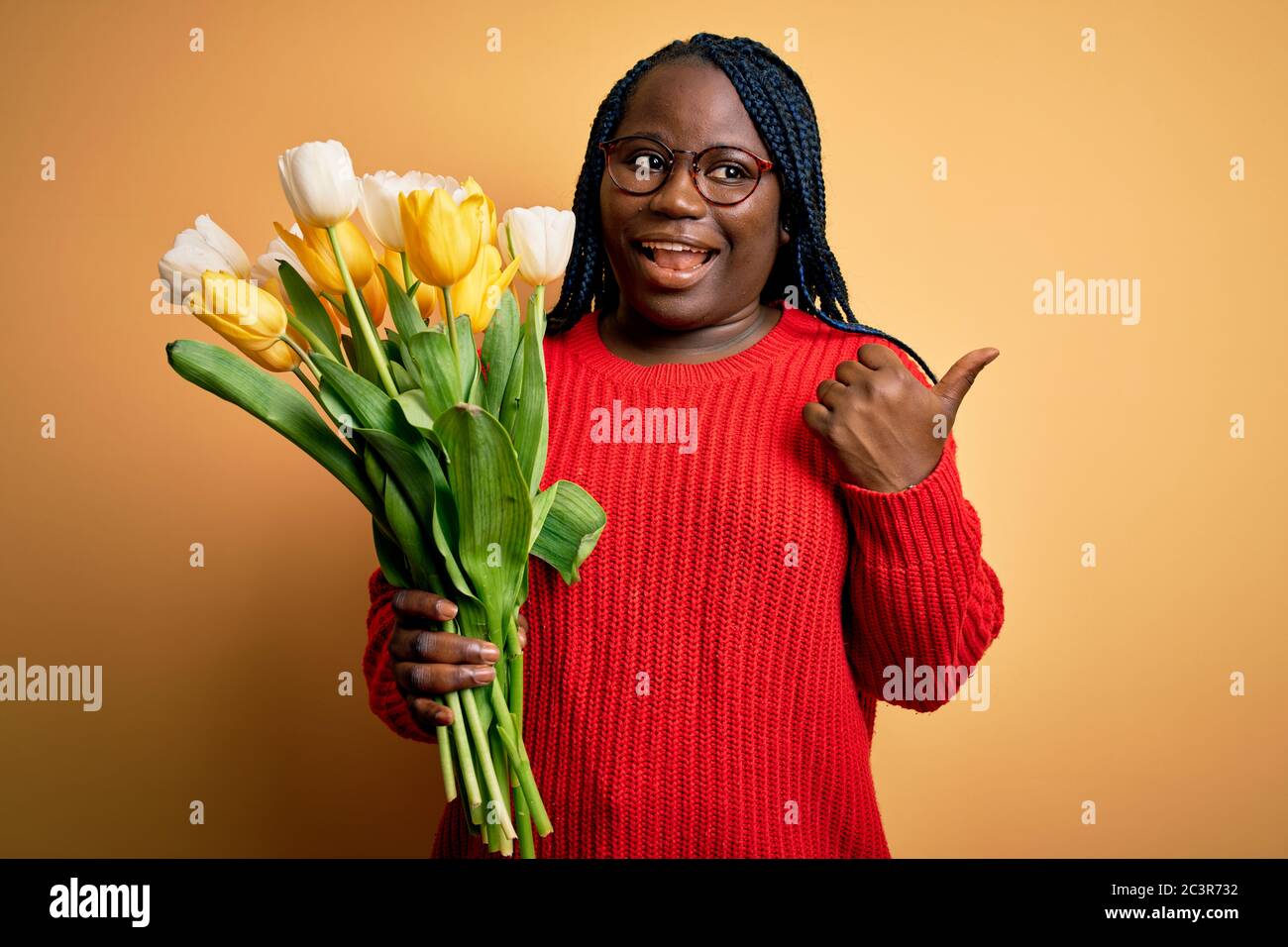 Young african american plus size woman with braids holding bouquet of yellow tulips flower smiling with happy face looking and pointing to the side wi Stock Photo