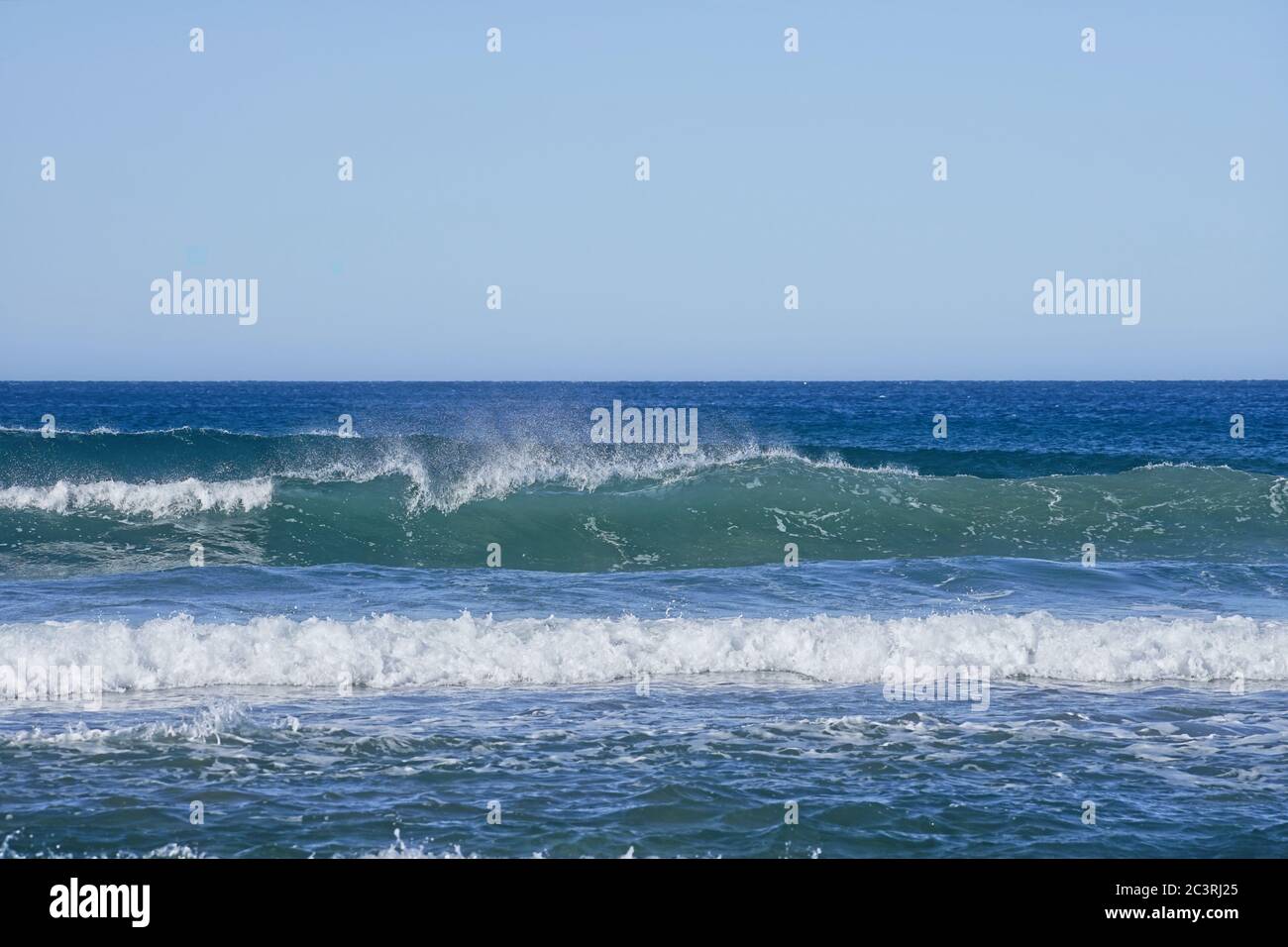 Sea wave breaking with distinctive spray blowing back from the top of the wave, blue sky above. Stock Photo