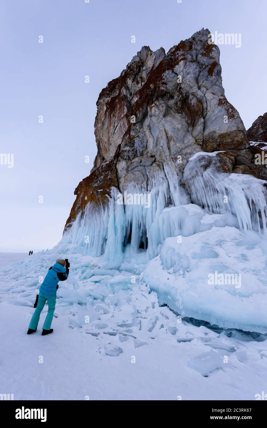 Lake Baikal, Russia - March 12, 2020: woman in a blue jacket takes photos of icy rocks with icicles on winter lake Baikal Stock Photo