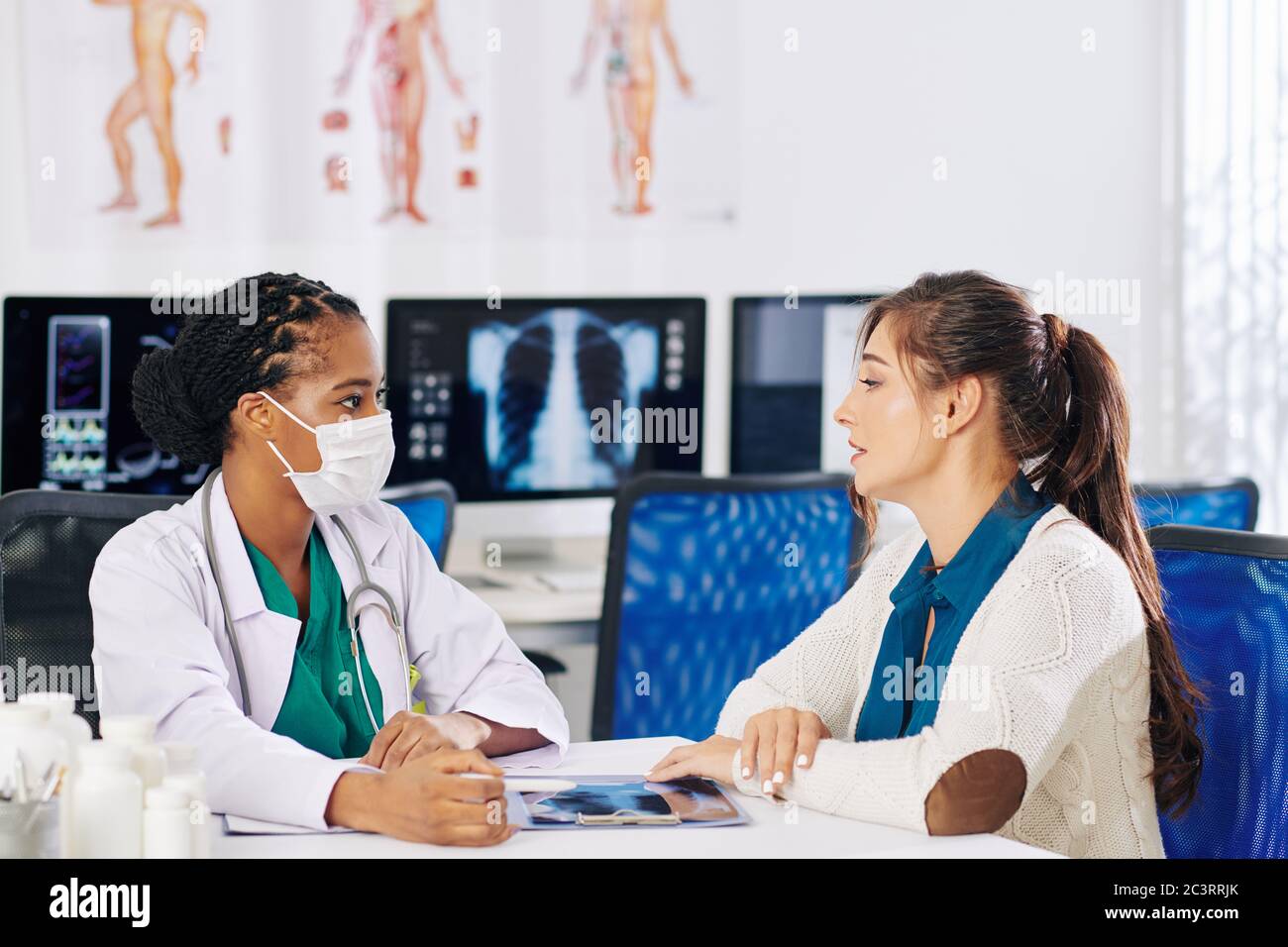 General practitioner in white labcoat and medical mask sitting at desk and talking to patient Stock Photo