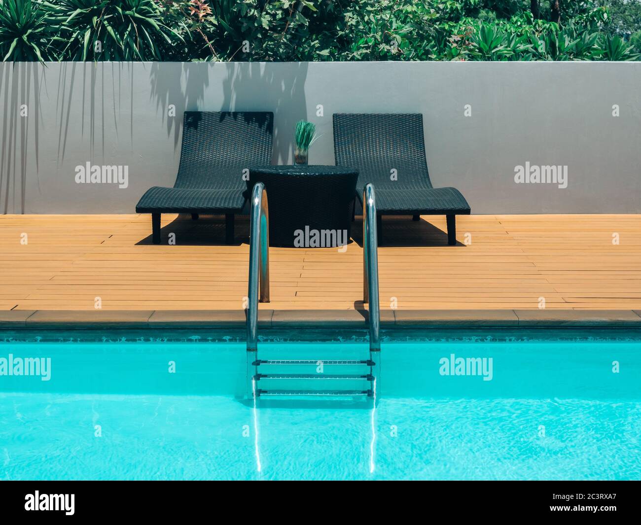 Outdoor swimming pool background minimal style. Grab bars ladder in the blue swimming pool with two black sunbed and green shrubs on grey wall. Stock Photo