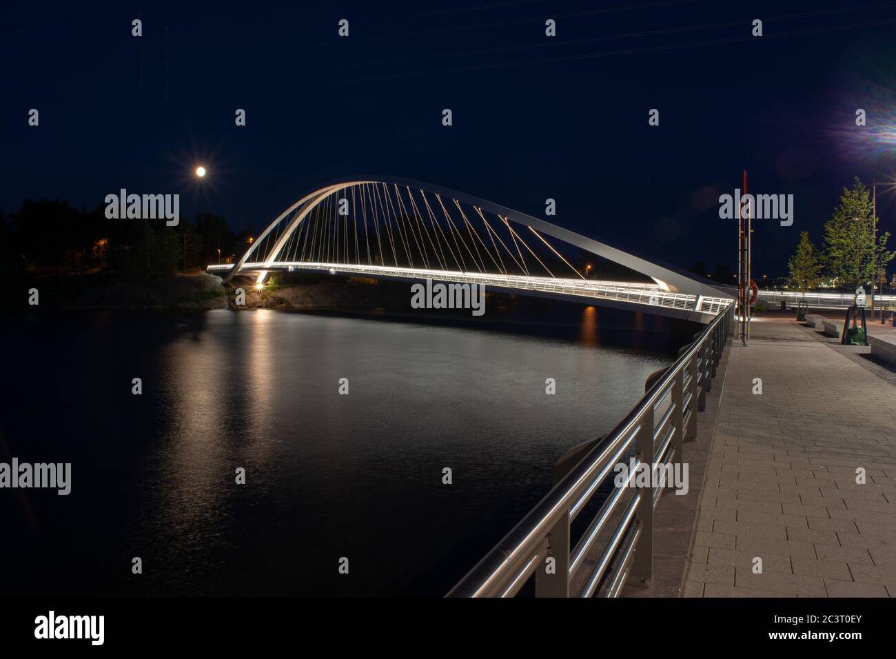 Illuminated arch type walking bridge reaches over the river at night. Stock Photo