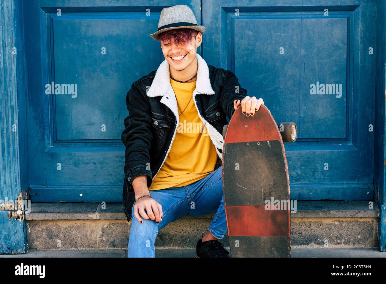 Cheerful and happy teenager sit down outside home with blue old door in background - city urban lifestyle and trendy generation z people concept - you Stock Photo
