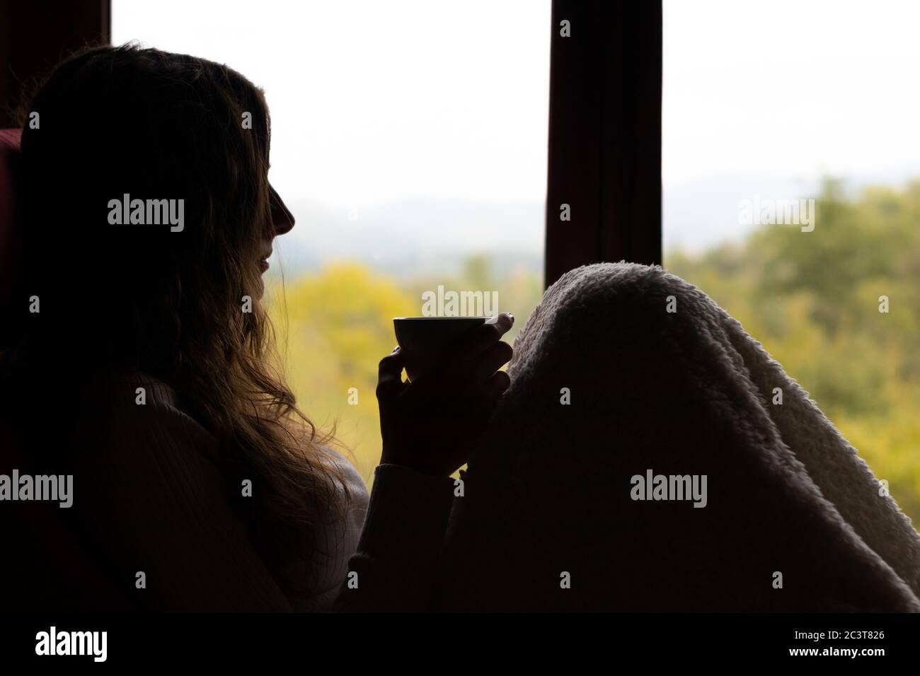 Silhouette of a young woman drinking a cup of coffee covered with a blanket while watching the landscape from a window. Stock Photo