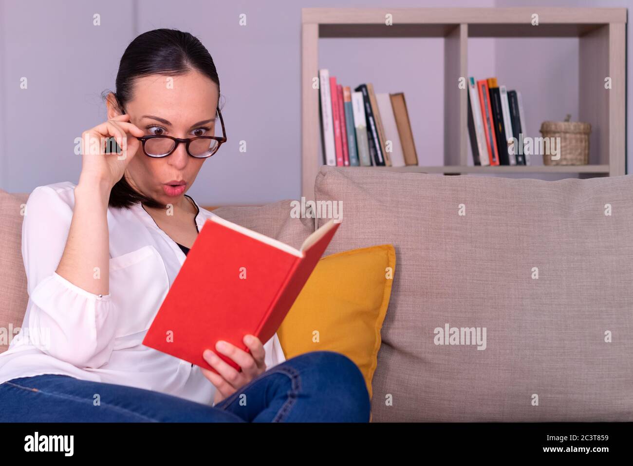 Pretty young brunette woman shocked reading a book sitting on the sofa at home. She is wearing a white shirt, glasses and a ponytail. Stock Photo