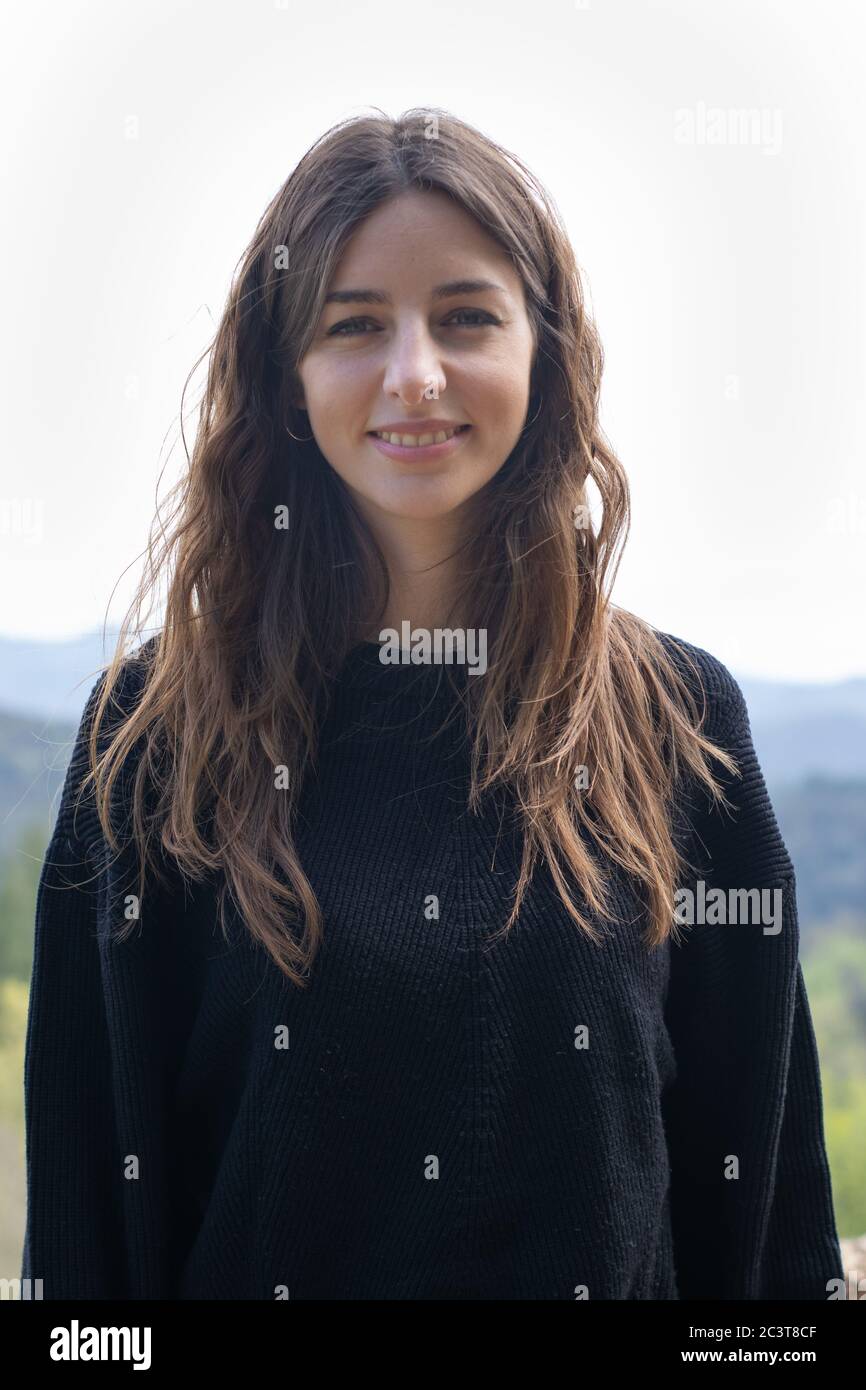Pretty young brunette woman smiling looking at camera standing. She is wearing a black sweater and her hair is down. Vertical image. Stock Photo
