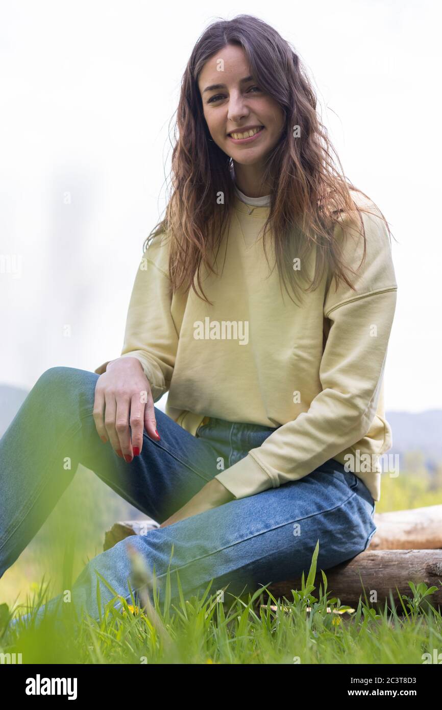 Pretty young brunette woman smiling and looking at camera sitting in a park outdoors on a sunny day. She is wearing blue jeans and a yellow sweater Stock Photo