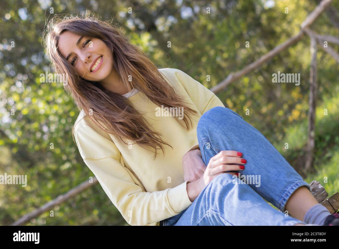 Pretty young brunette woman smiling and looking at camera sitting in a park outdoors on a sunny day. She is wearing blue jeans and a yellow sweater Stock Photo