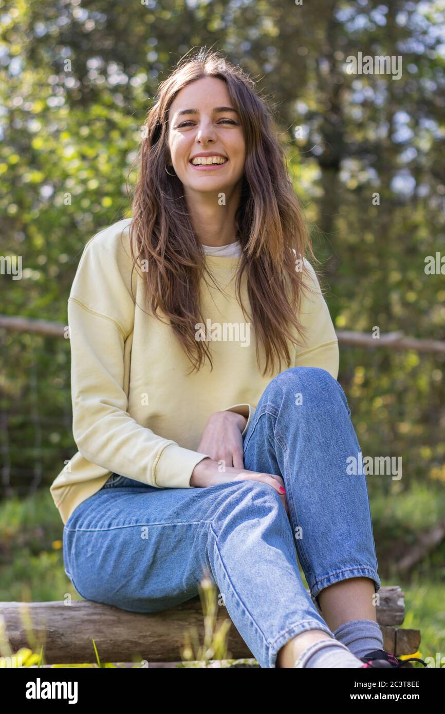 Pretty young brunette woman laughing and looking at camera sitting in a park outdoors on a sunny day. She is wearing blue jeans and a yellow sweater Stock Photo