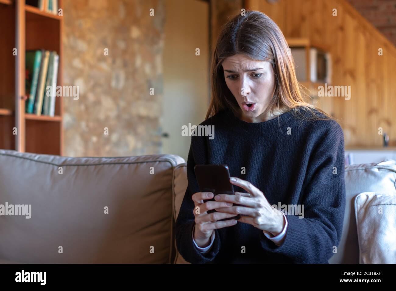 Shocked pretty young brunette woman typing on the smartphone sitting on the sofa at home wearing black sweater Stock Photo