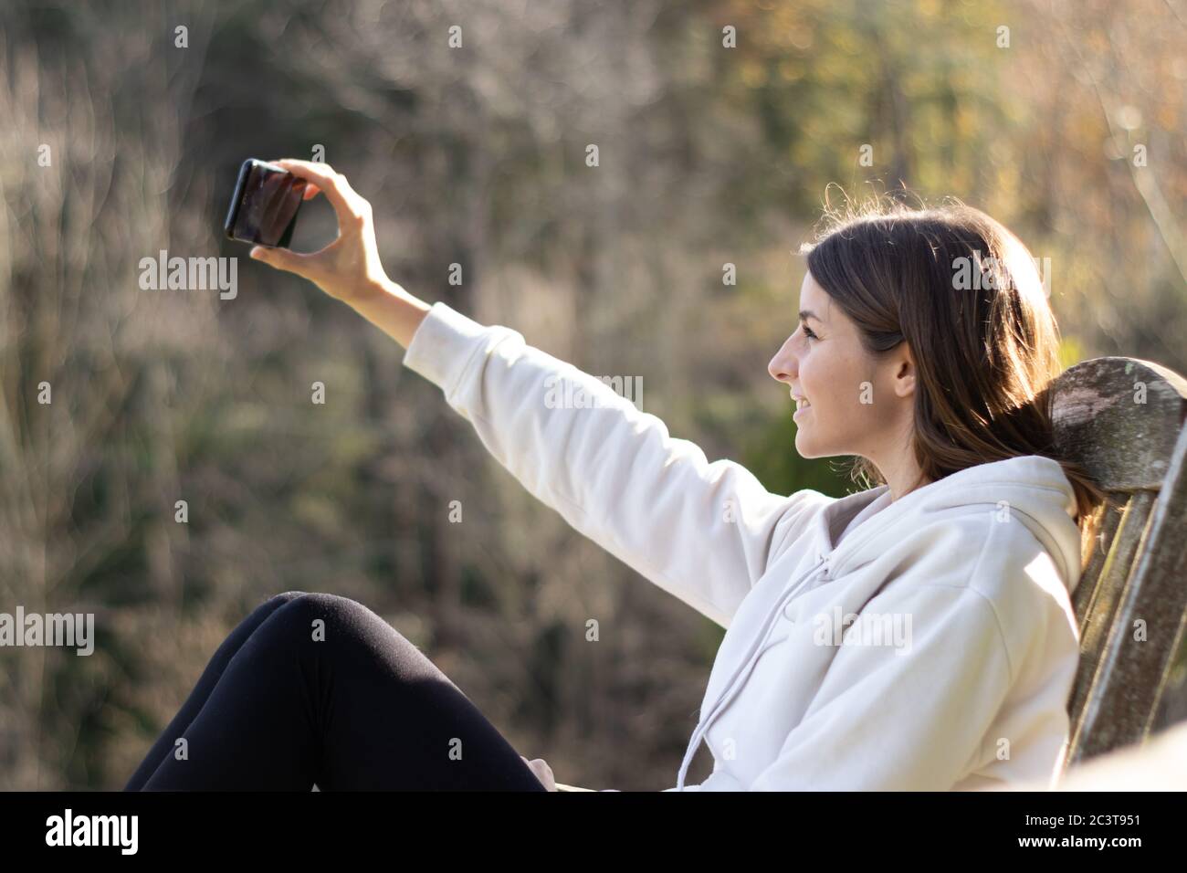 Beautiful smiling brunette woman takes a selfie with the smartphone sitting in the park on a sunny day. Stock Photo