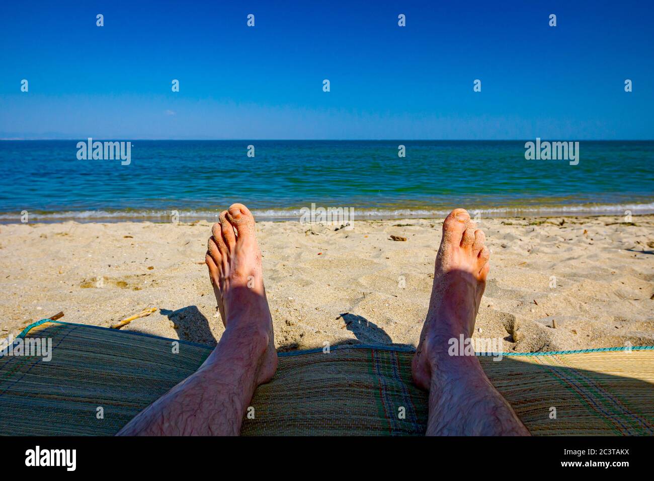 Man's legs are sunbathing by lying carefree on mat next to the coastline, on public beach. Stock Photo