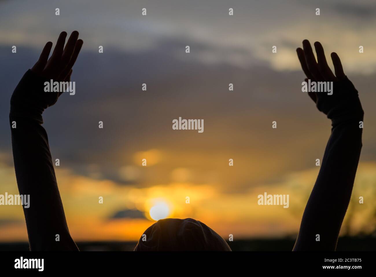 Person raising their hands silhouetted against a vivid orange sunset with the orb of the sun touching the top of their head Stock Photo