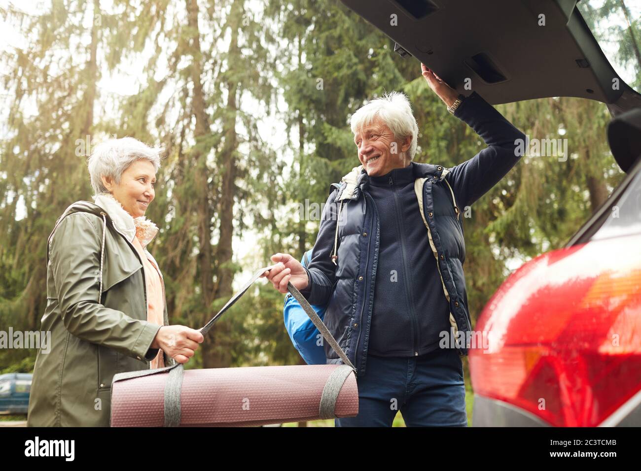 Low angle portrait of modern senior couple opening trunk of car and smiling happily while going on hike in forest Stock Photo