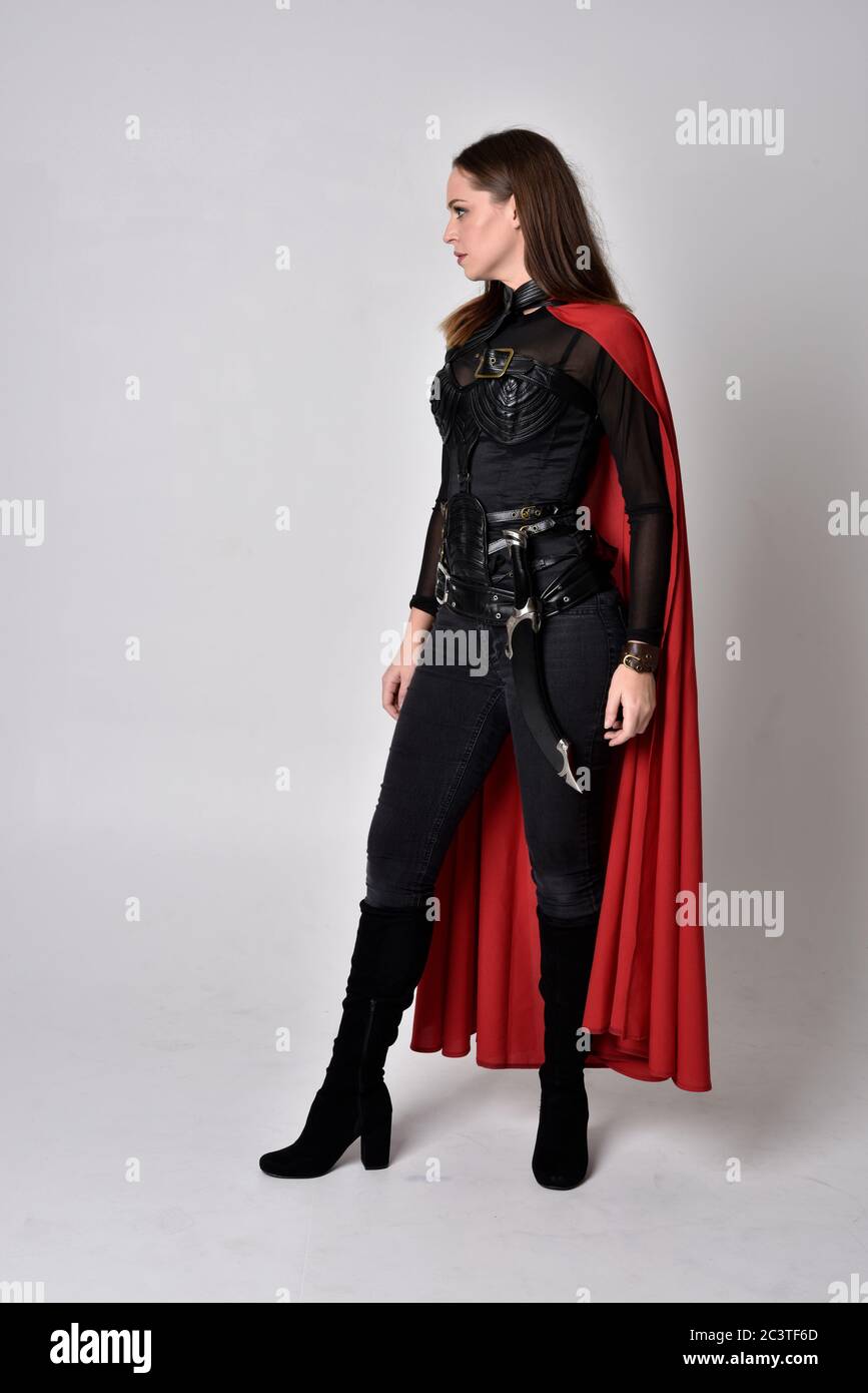 Portrait of brunette girl wearing black leather catsuit & red cape.  full length standing pose, isolated against a studio background. Stock Photo