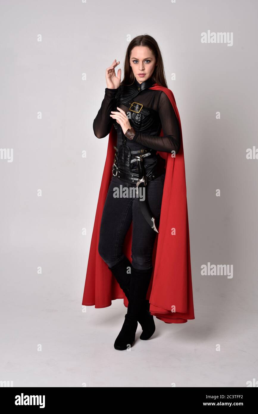Portrait of brunette girl wearing black leather catsuit & red cape.  full length standing pose, isolated against a studio background. Stock Photo