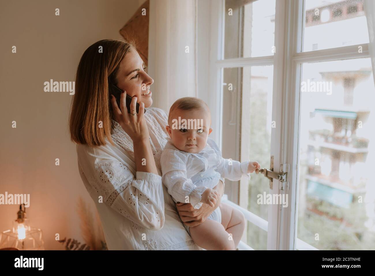 Young mother talks on the phone looking out the window with her baby in arms Stock Photo