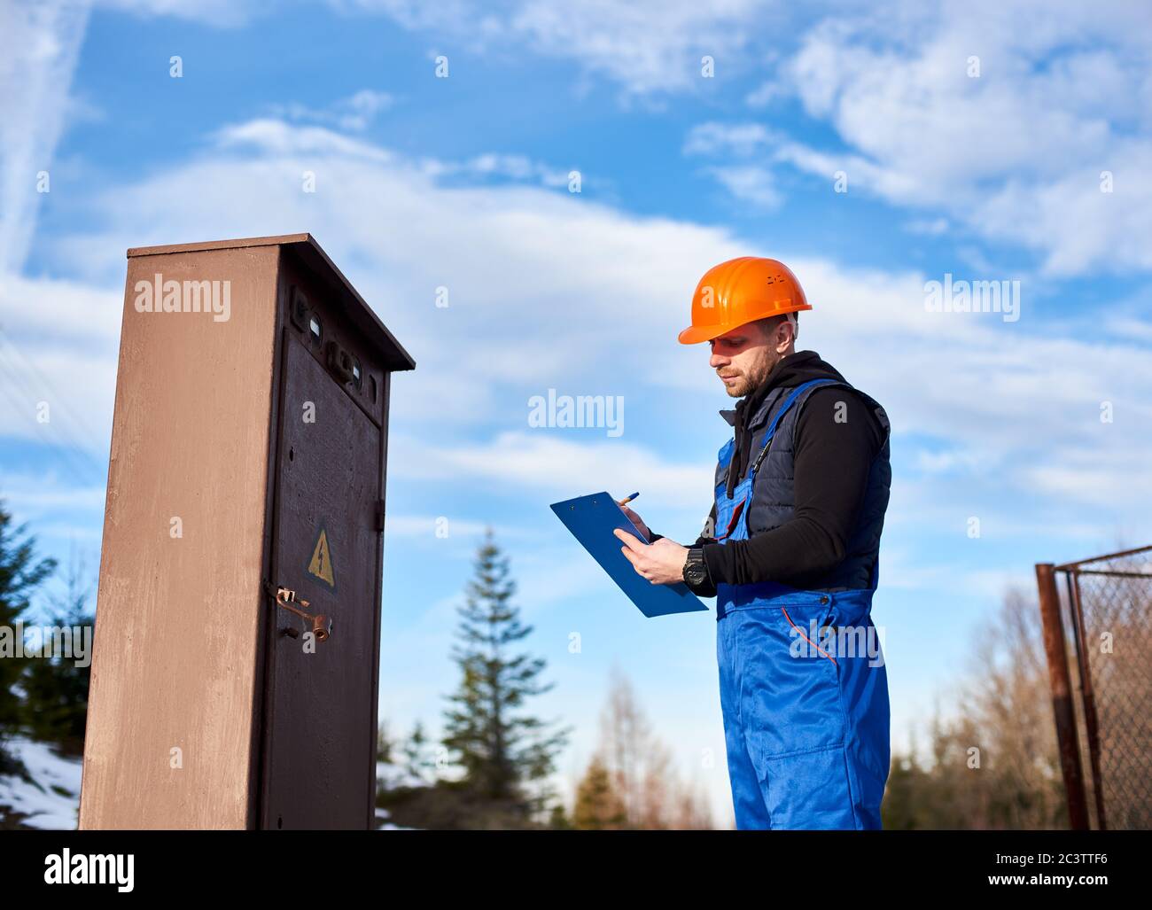 Oil inspector in protective overalls and orange helmet making notes next to a transformer outdoors against blue sky with white clouds. Concept of petroleum industry. Stock Photo