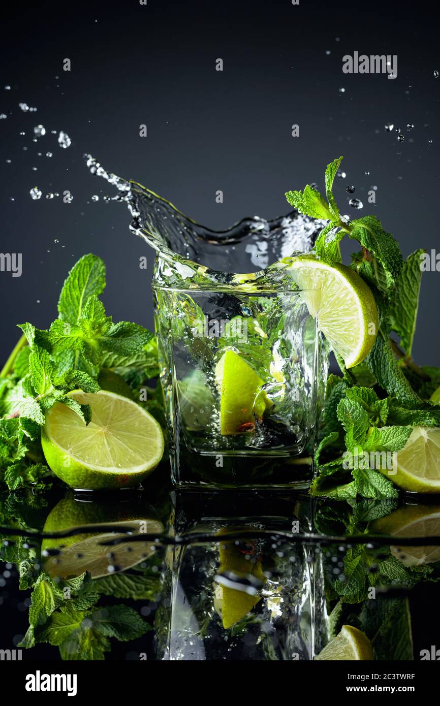 Cocktail with limes and mint on a black reflective background. Lime slice fall in glass with drink. Stock Photo