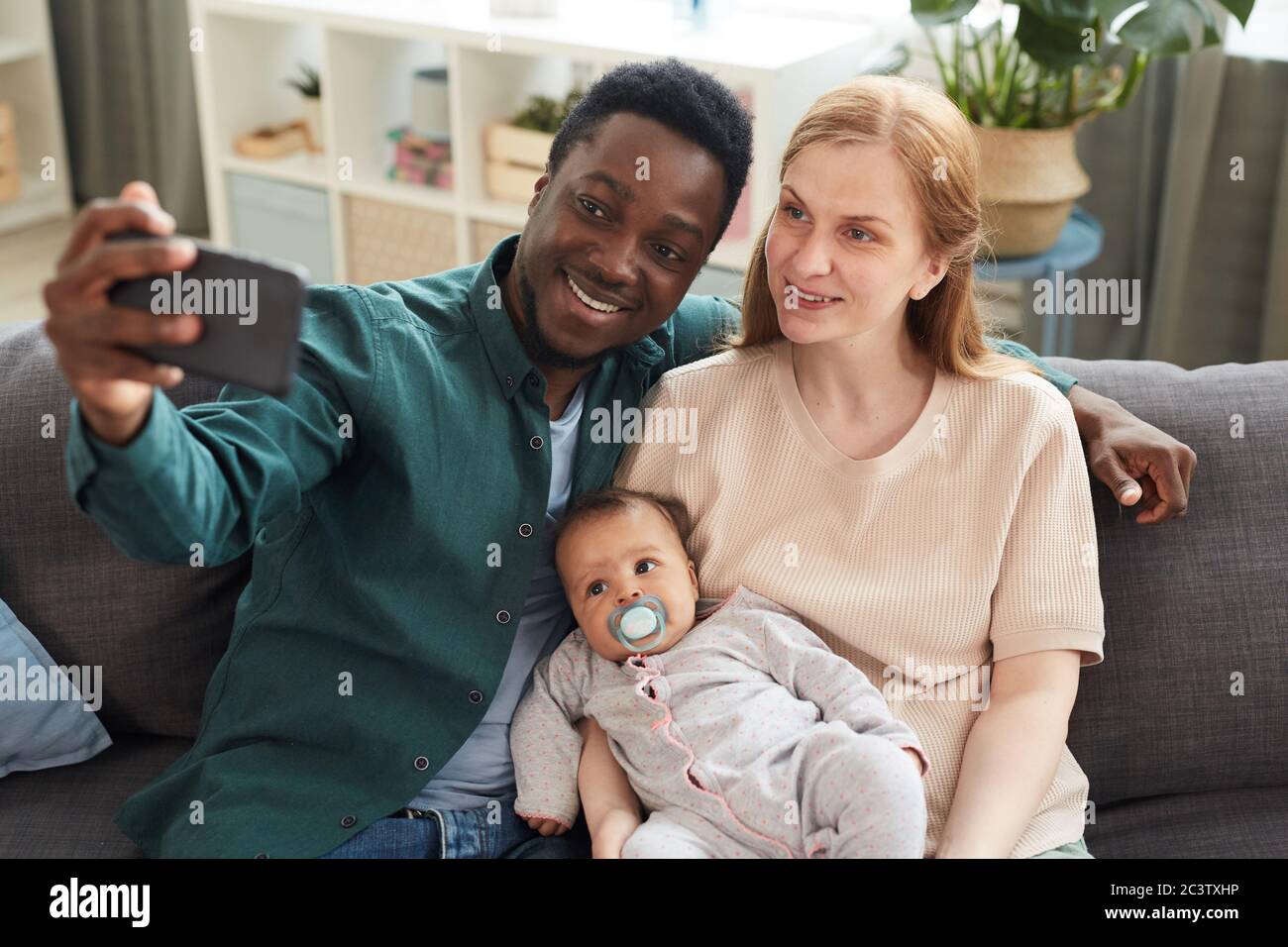 High angle portrait of young interracial family taking selfie photo with cute mixed-race baby while sitting on couch in home interior Stock Photo