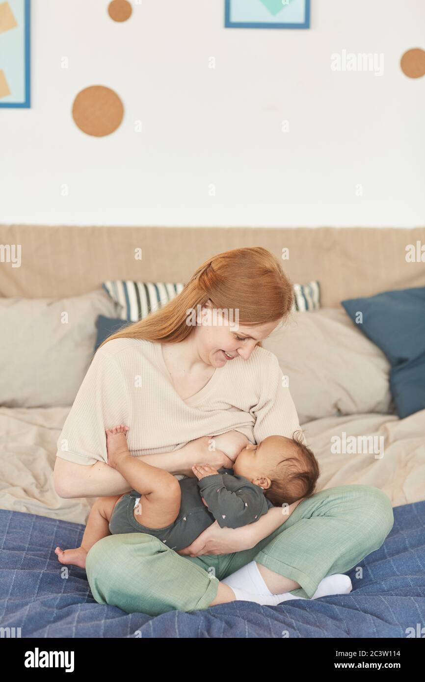 Vertical full length portrait of smiling adult mother breastfeeding mixedrace baby while