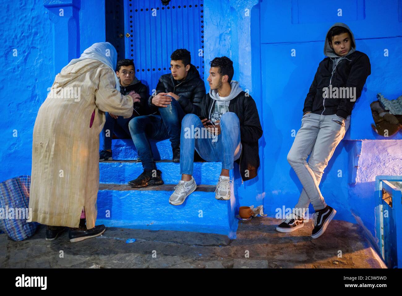 Morocco: Chefchaouen, the blue city. Atmosphere in the Old Town: old woman in traditional clothes talking with young men sitting on a staircase in a l Stock Photo