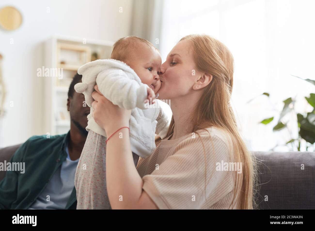 Portrait of happy interracial family at home, focus on Caucasian woman kissing cute baby in foreground, copy space Stock Photo