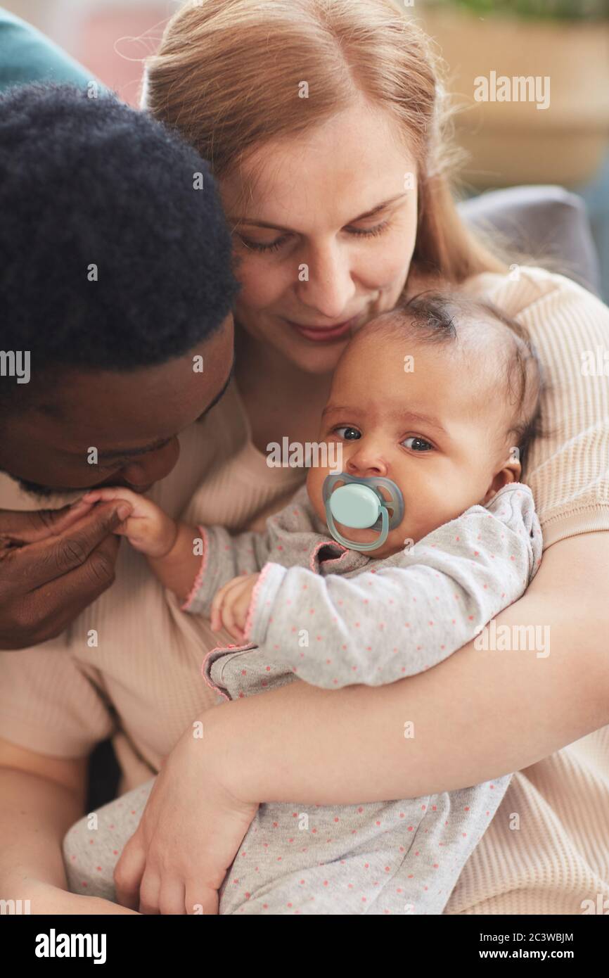 Vertical warm-toned portrait of loving interracial family holding baby boy, with African-American father gently kissing tiny hand Stock Photo