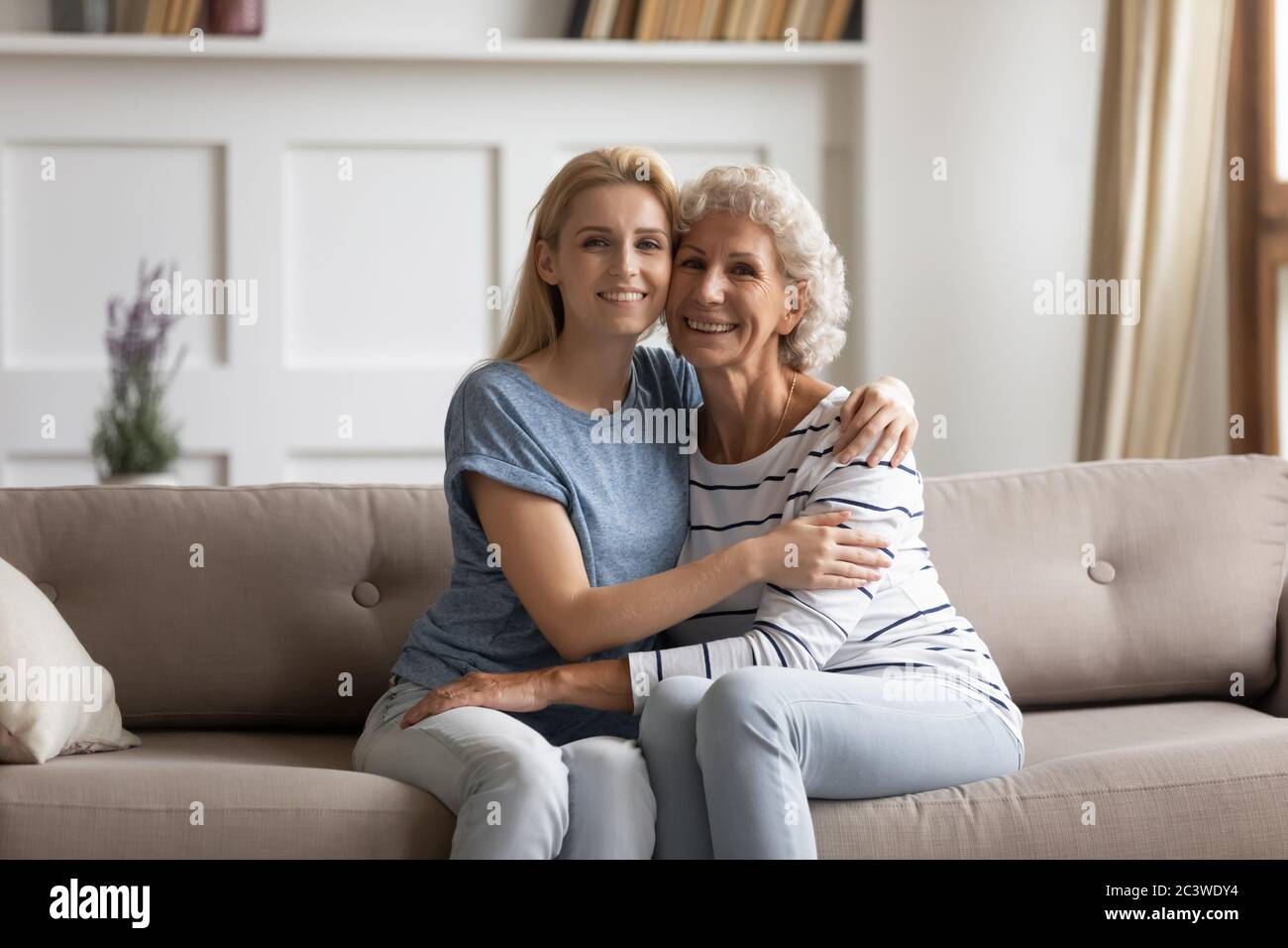Senior mother hugs young grownup daughter sitting together on couch Stock Photo
