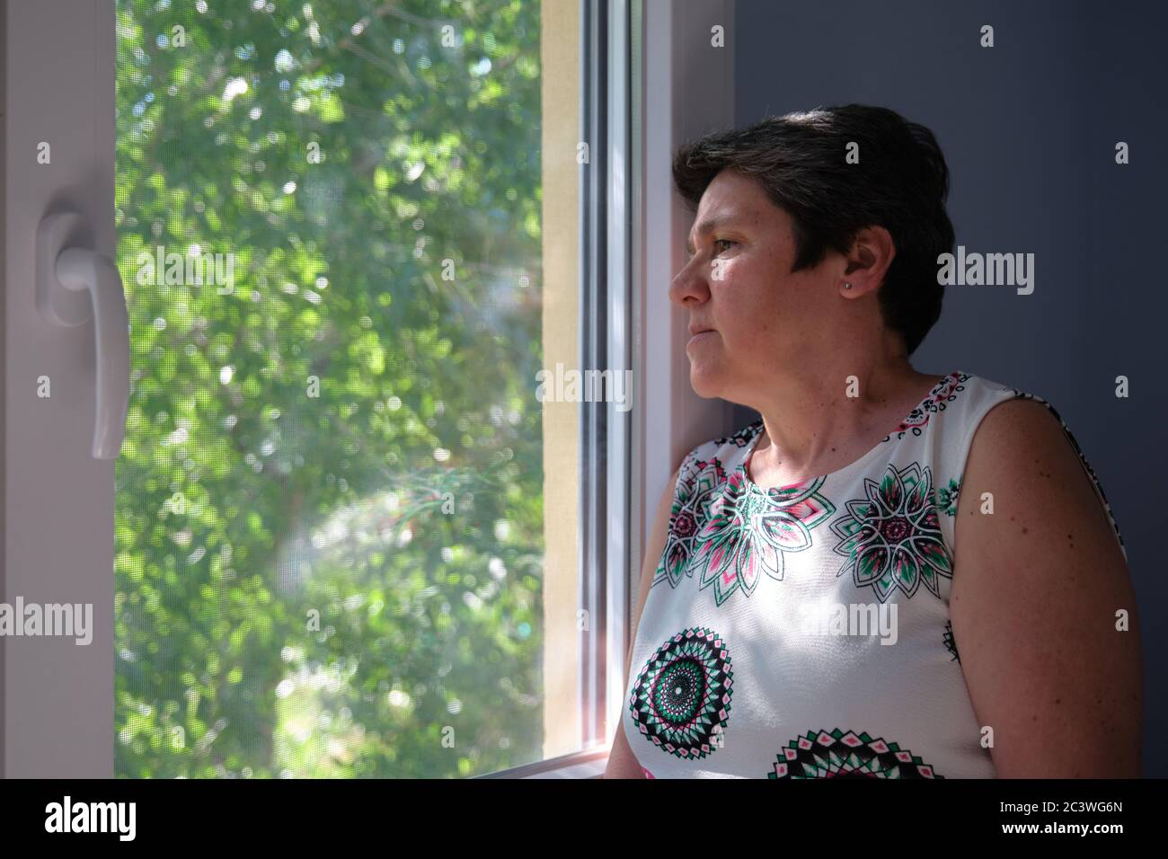 Sad, worried, thoughtful mature woman looking through the window. Sadness and quarantine concept. Stock Photo