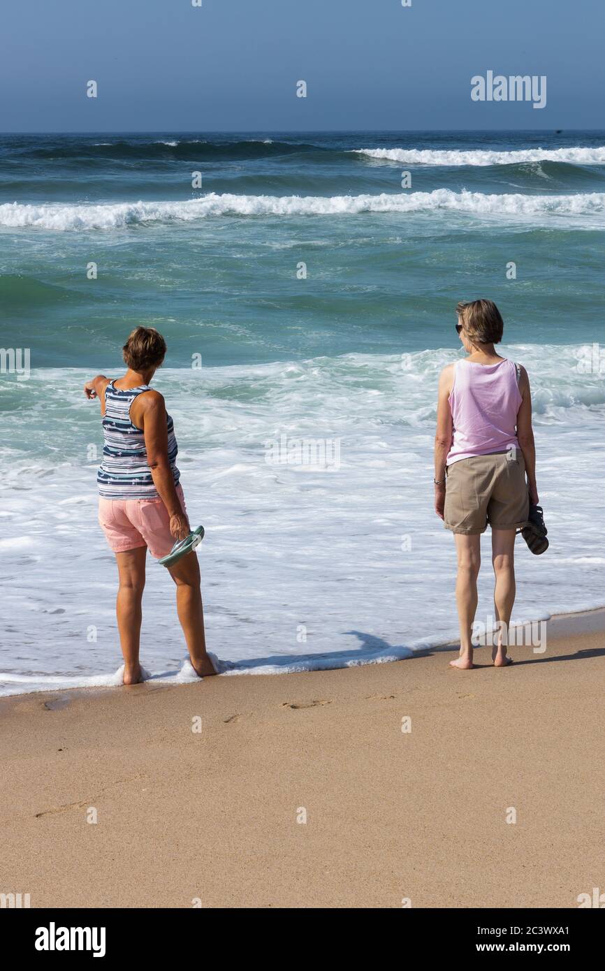 Two relaxed looking senior woman on beach walking at edge of sea with white waves rolling. Portugal Stock Photo