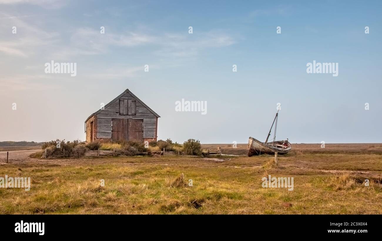 The old coal barn at Thornham harbour North Norfolk  coast with old abandoned disused fishing boat lying to one side in the spring evening sun Stock Photo