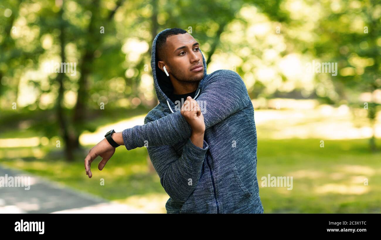 Athletic African American guy in hoodie stretching his arm and listening to music after training at summer park Stock Photo
