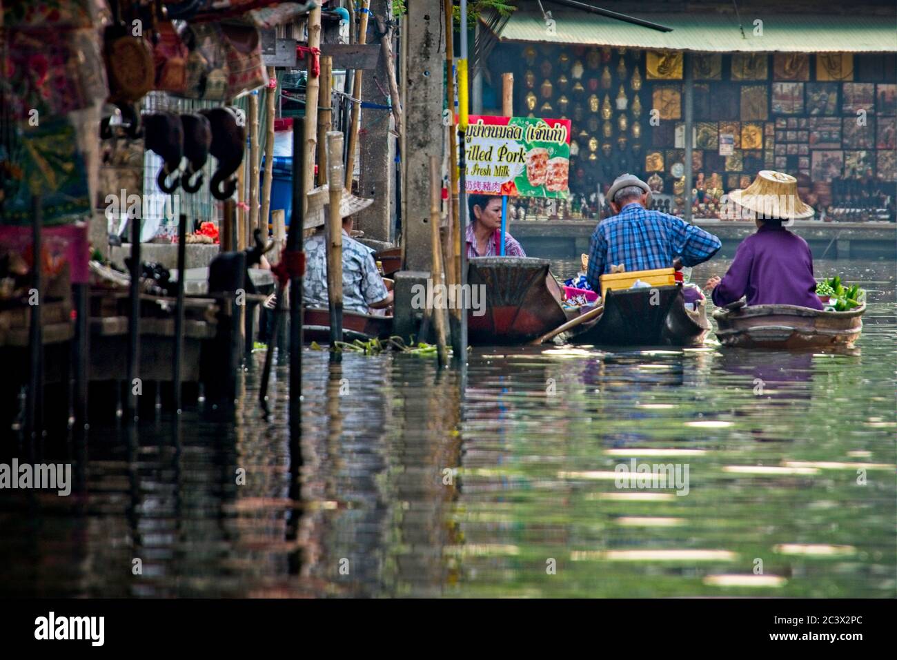 Locals at Bangkok's floating market Rear view of four boats with people Reflections in water Goods for sale in shop on left and background Landscape Stock Photo