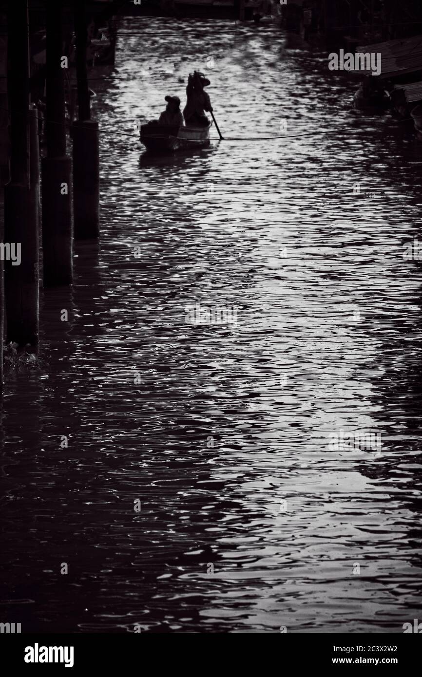 Bangkok's floating market Mono process Single boat in distance with two people Silhouetted against highlight in water Stock Photo